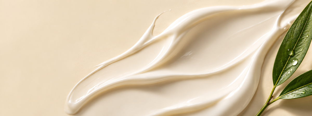 Smooth white moisturising cream spread across a warm beige background with a green leaf and water droplets.